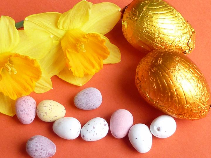 daffodils_eggs_background.jpg - Yellow daffodils, colorful quail egg candies and chocolate eggs wrapped in golden foil, viewed from above in close-up on orange surface background