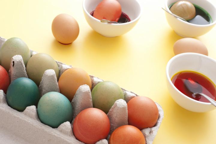 easter_colouring_eggs.jpg - Slanted view of carton with a dozen colorful easter eggs beside three bowls of dye