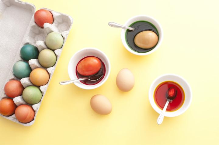 making_coloured_eggs.jpg - From above view of three bowls with paint, spoons and eggs next to colorful eggs in the box on yellow background