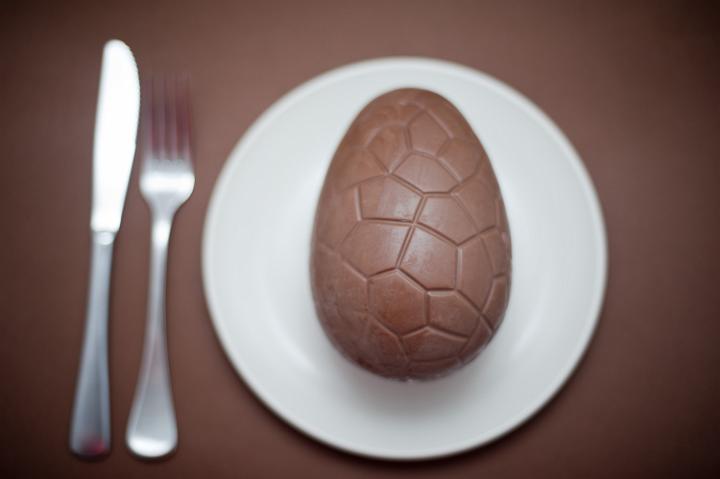 concept_easter_egg.jpg - Top view of chocolate patterned easter egg on white plate next to silver fork and knife on brown background.