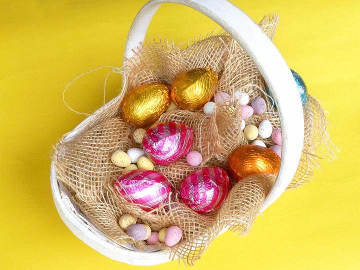 easter_collecting_basket.jpg - White wicker basket for collecting Easter eggs, with various colorful chocolate egg treats on sack cloth, viewed from above on yellow background