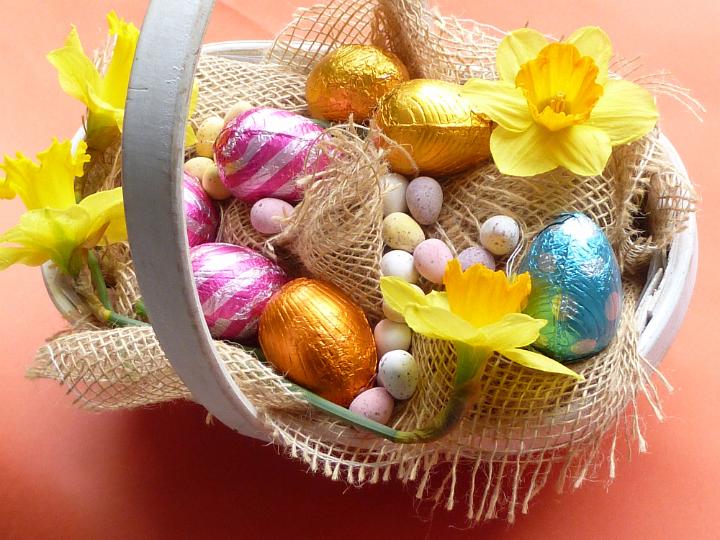 easter_eggs_in_bakset.jpg - Beautiful rustic basket with chocolate Easter eggs, decorated with sack cloth and fresh yellow narcissus or daffodil flowers, viewed in close-up from above on red background